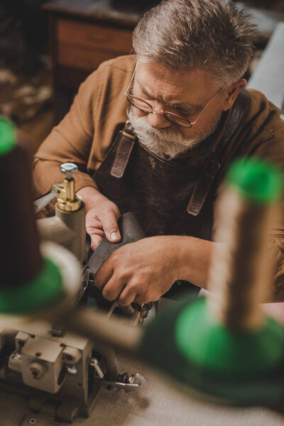 selective focus of senior shoemaker sewing leather on sewing machine in workshop