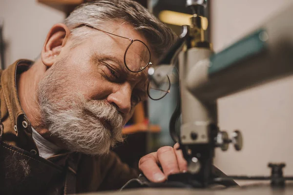 selective focus of bearded, senior cobbler sewing leather on sewing machine in workshop