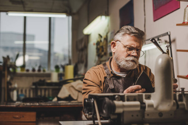 senior, bearded cobbler sewing leather on sewing machine in workshop
