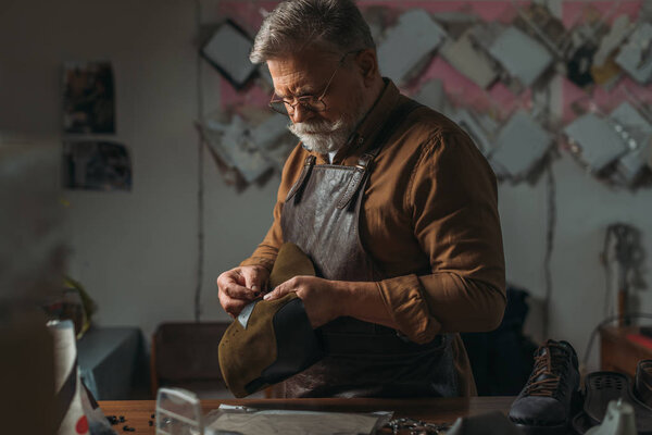 selective focus of senior cobbler holding piece of genuine leather in workshop