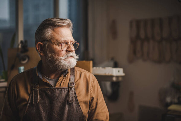 confident, senior cobbler in glasses looking away while standing in workshop