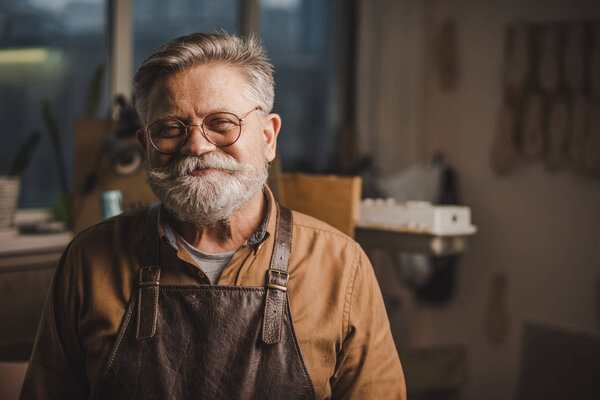 happy, senior shoemaker in glasses smiling at camera while standing in workshop