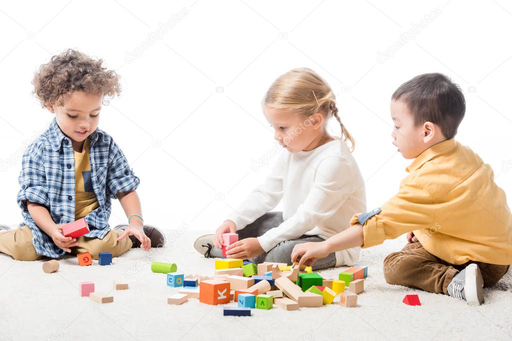 multicultural kids playing with wooden blocks on carpet, isolated on white