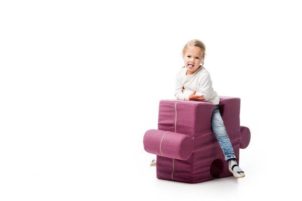 happy kid sticking tongue out while sitting on purple puzzle chair, isolated on white 