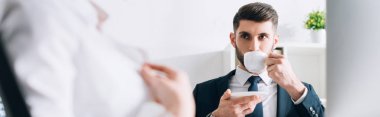 panoramic shot of businessman drinking coffee and looking at secretary in office 