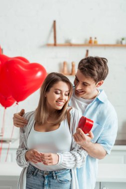 Balonların yanında kalp şeklinde hediye kutusu tutan bir adam ve büyük göğüslü mutlu bir kız arkadaş. 