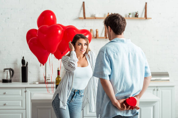 back view of man holding heart-shaped gift box near attractive girlfriend at home 