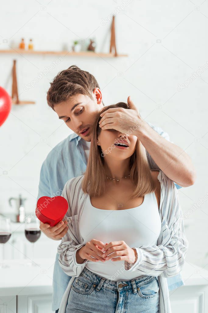 man holding heart-shaped gift box and covering eyes of girlfriend with big breast 