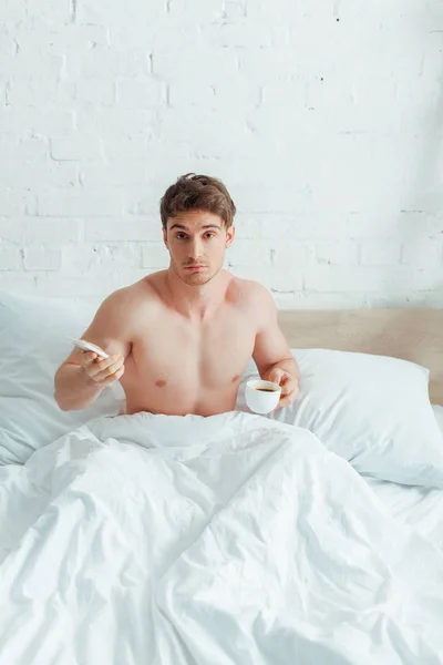 Förvirrad Man Som Håller Kopp Kaffe Och Smartphone Sängen — Stockfoto