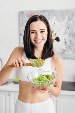 Beautiful sportswoman smiling at camera while holding bowl with fresh salad in kitchen