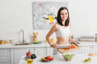 Attractive girl smiling at camera while cooking salad with raw vegetables in kitchen