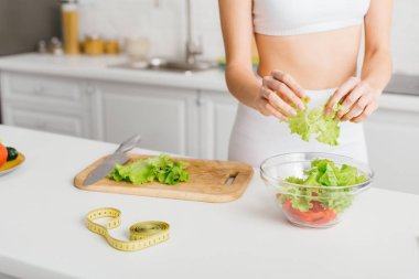 Cropped view of fit girl preparing fresh salad with avocado near measuring tape on kitchen table  