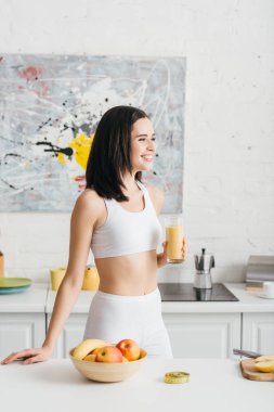 Meyvelerin yanında gülümseyen ve mutfak masasındaki şeritleri ölçen bir bardak smoothie ile zayıf bir sporcu kadın.