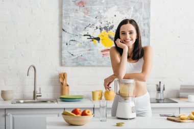 Slim sportswoman smiling at camera while preparing smoothie near measuring tape on kitchen table