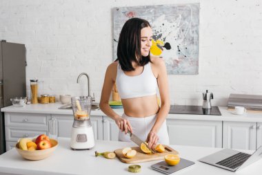 Smiling fit sportswoman looking at laptop while cutting fruits near blender on kitchen table, calorie counting diet