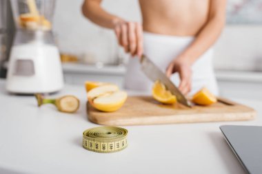 Selective focus of measuring tape and scales near fit girl cutting fresh vegetables for smoothie on kitchen table, calorie counting diet clipart