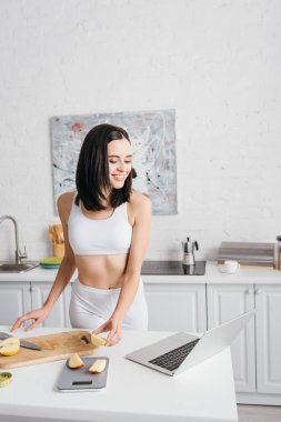 Beautiful smiling sportswoman looking at laptop while cutting apple near scales and measuring tape on kitchen table, calorie counting diet