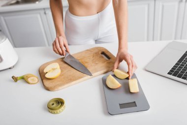 Cropped view of slim girl cutting fresh fruits near scales, measuring tape and laptop on kitchen table, calorie counting diet