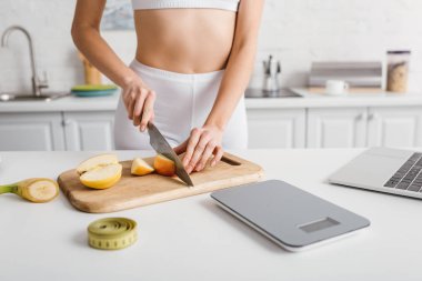 Cropped view of sportswoman cutting fruits near laptop, scales and measuring tape on kitchen table, calorie counting diet
