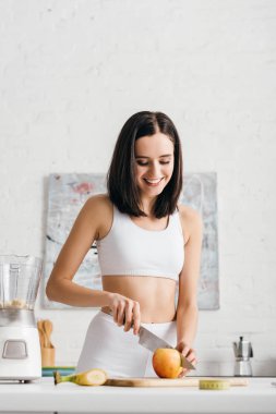 Selective focus of beautiful smiling sportswoman preparing smoothie with fruits near measuring tape on table