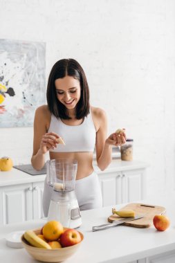 Beautiful smiling sportswoman putting banana in blender while preparing smoothie 