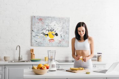 Beautiful smiling sportswoman preparing smoothie near laptop and measuring tape in kitchen