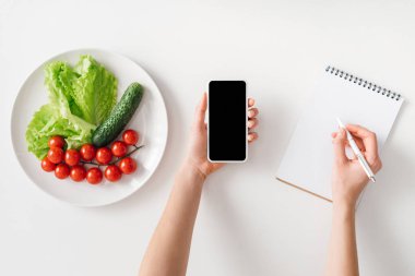 Top view of woman holding smartphone and writing in notebook near ripe vegetables on plate on white background