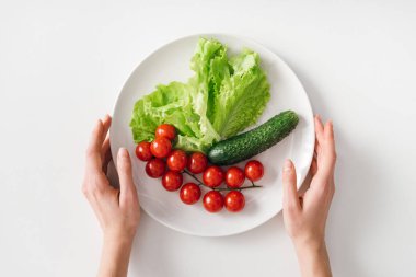 Top view of woman holding plate with raw vegetables on white background
