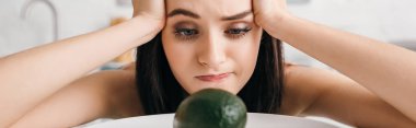 Selective focus of pensive girl with hands near head looking at avocado on kitchen table, panoramic shot 