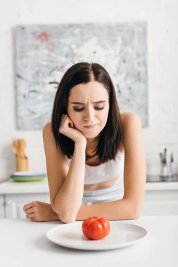 Pensive sportswoman looking at ripe tomato on plate on table