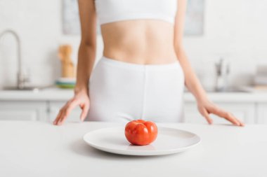 Selective focus of ripe tomato on plate and slim sportswoman near kitchen table