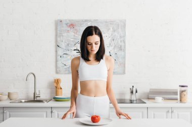 Beautiful sportswoman looking at tomato on kitchen table