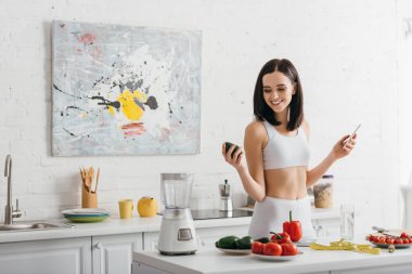 Smiling sportswoman holding avocado and smartphone near vegetables and measuring tape on table, calorie counting diet
