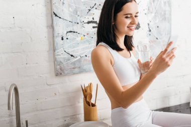 Gülümseyen sporcu kadın mutfak masasında bir bardak su ve akıllı telefon tutuyor.