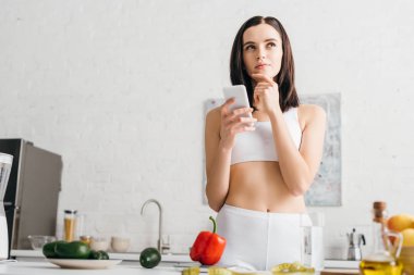 Pensive sportswoman using smartphone near fresh vegetables, fruits and measuring tape on table