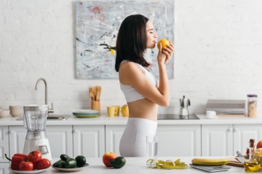 Side view of fit sportswoman holding orange near fruits, vegetables and measuring tape on kitchen table, calorie counting diet