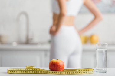 Selective focus of apple, measuring tape and glass of water near fit sportswoman in kitchen