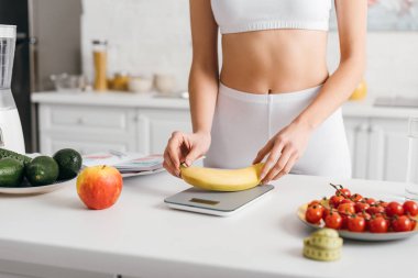 Selective focus of fit sportswoman putting banana on scales near measuring tape and notebook on table, calorie counting diet