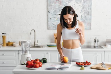 Smiling sportswoman weighing apple near notebook and measuring tape on kitchen table, calorie counting diet