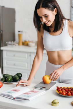 Selective focus of beautiful smiling sportswoman writing calories while weighing apple on kitchen table, calorie counting diet