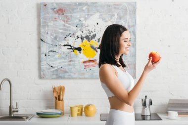 Side view of smiling sportswoman holding fresh apple in kitchen