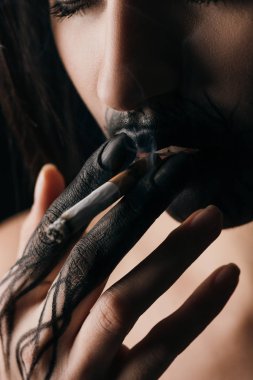 Selective focus of girl with black painted hand and lips smoking cigarette isolated on black 