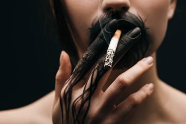 Selective focus of woman with black painted hand and lips smoking cigarette isolated on black  clipart