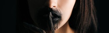 Panoramic shot of woman with black painted lips and hand showing quiet gesture isolated on black 