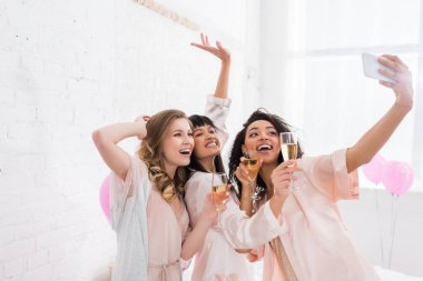 Heyecanlı çok kültürlü kızlar ellerinde şampanya kadehleriyle pijama partisinde akıllı telefondan selfie çekiyorlar.