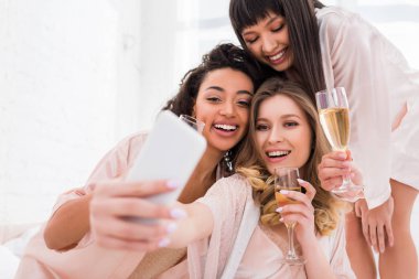 Gülümseyen, şampanya kadehli kızlar pijama partisi sırasında akıllı telefondan selfie çekiyorlar.