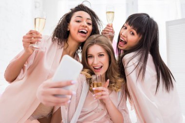 Şampanyalı neşeli çok kültürlü kızlar pijama partisi sırasında akıllı telefondan selfie çekiyorlar.