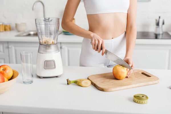 Cropped view of sportswoman cutting fruits for smoothie near measuring tape on table