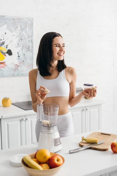 Smiling Fit Sportswoman Holding Banana Pieces While Preparing Smoothie Kitchen — Stockfoto