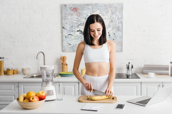 Smiling sportswoman preparing smoothie near laptop, smartphone and notebook on table in kitchen